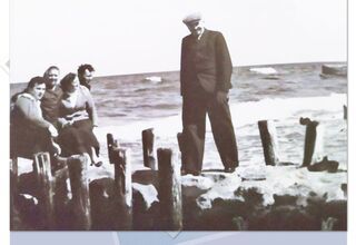 Plakat Spotkania Klubu Historycznego. Starszy pan stoi w otoczeniu siedziących kobiet nad brzegiem morza
