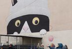 Mural z głową kota i wizerunkiem babci robiącej na drutach, przed muralem stoją ludzie