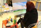 Kobieta  siedzi na krześle i  maluje obraz na sztaludze