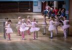 Dziewczynki w różowych strojach baletowych trzymają się drążka, obok dorośli na krzesłach